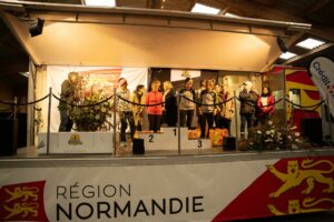 Vue large de la remise des récompenses, drapé Région Normandie au premier plan : lauréates et organisateurs autour du podium, lots posés au sol.