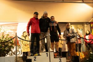 Trois coureurs bras dessus bras dessous sur les marches du podium, un officiel présentant la coupe à droite.