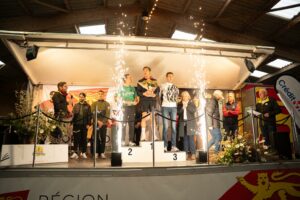 Podium masculin sous les étincelles, le speaker au micro à gauche : le vainqueur au centre avec son bouquet.