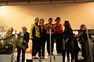Podium féminin : les lauréates médaillées sur les marches, bouquet à la main, félicitées par les organisatrices.