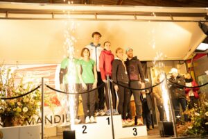 Plusieurs binômes de coureurs réunis sur les trois marches du podium, sous les étincelles.
