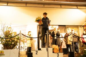 Le vainqueur seul sur la marche 1, bouquet et trophée à la main, applaudi par les officiels.